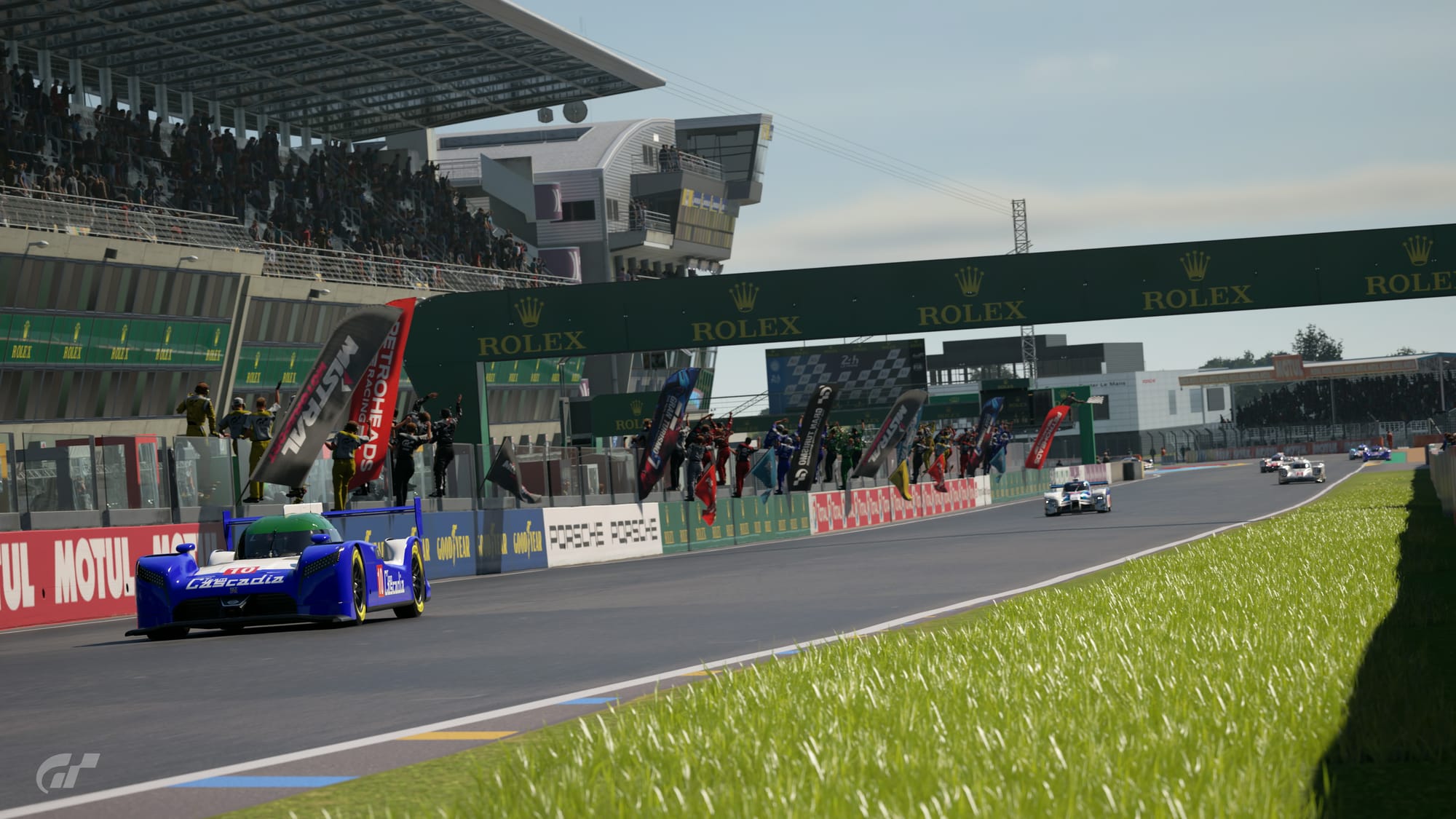 Six Le Mans Prototype race cars on the front straight at Circuit De La Sarthe as the race has just finished, flag man waving the chequered flag and team crew members celebrating up on the pit wall. The car closest in shot is a blue, with white and green Nissan GT-R LM Nismo that says Team Cascadia on the front.