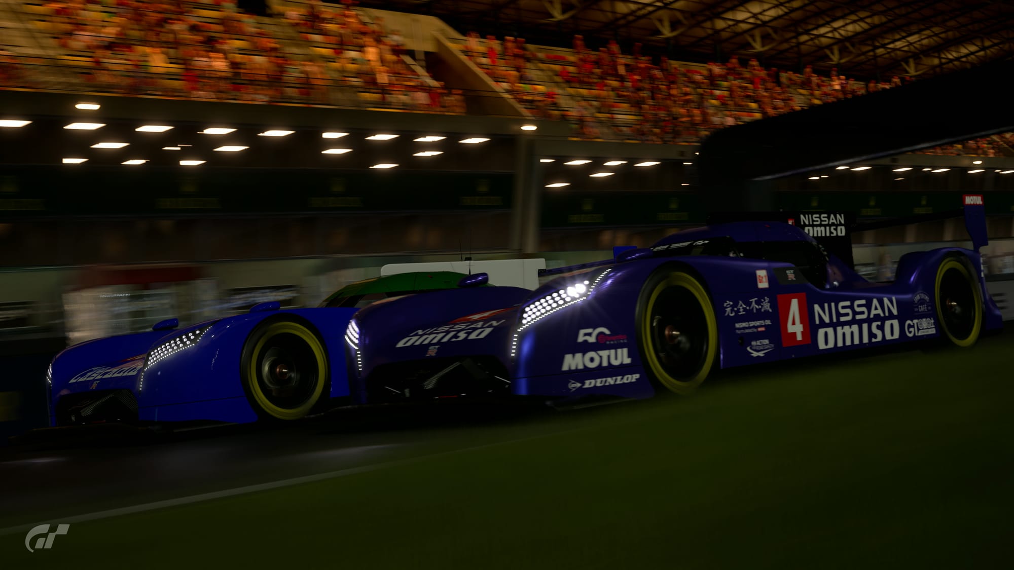 Two blue Nissan GTR LM NISMO racecars go side by side on the front straight at Circuit De La Sarthe, with the far car starting to nose ahead.