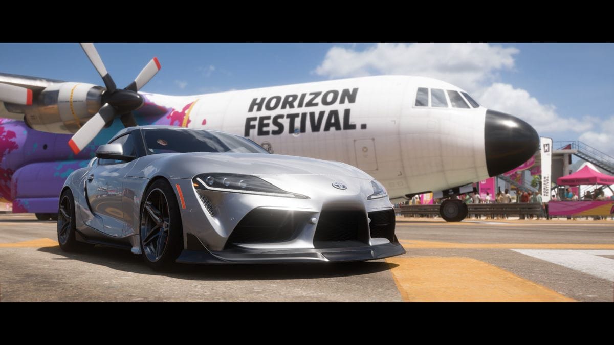 A Silver A90 Toyota Supra with too big of wheels is parked in front of a turbo-prop plane that's on a taxiway. The plane says Horizon Festival. on the side. It is a screenshot from Forza Horizon 5.