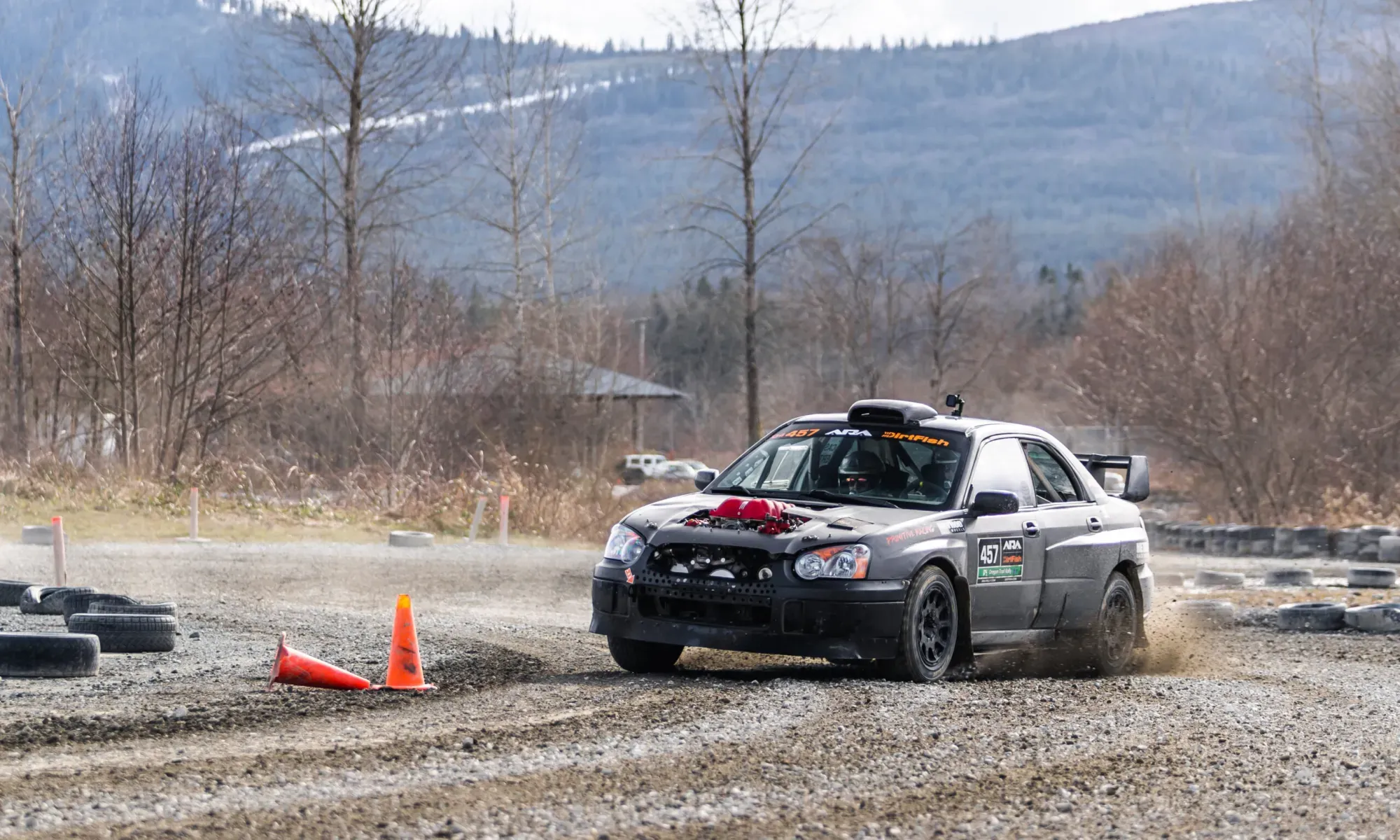 A grey Subaru Impreza rally car going through a gravel course. It has a section of it's hood cut out, revealing the red intake manifold normally seen on a Ferrari V8.
