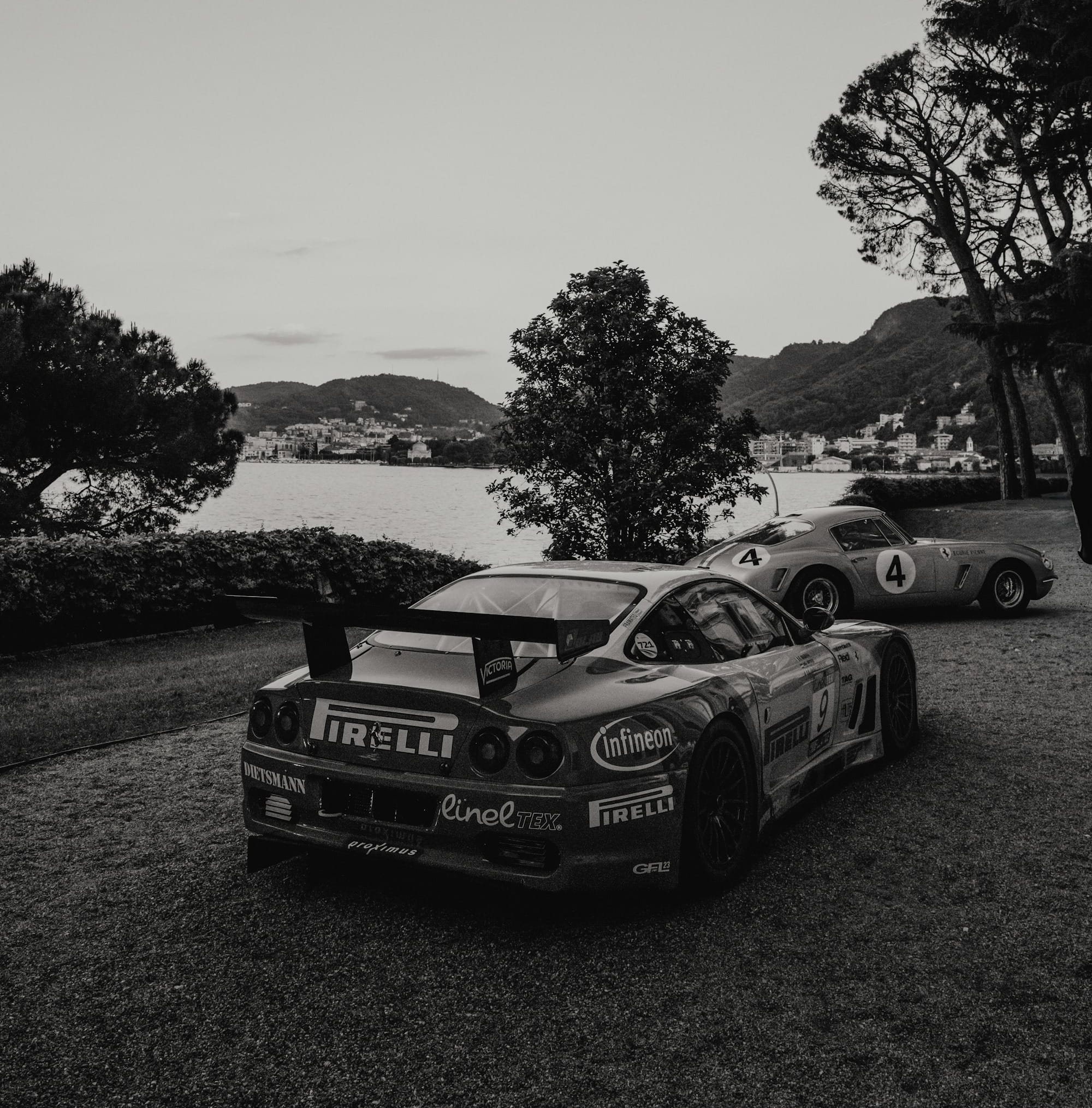 A Ferrari 575 GTC and a Ferrari 375 parked in a yard overlooking a lake.