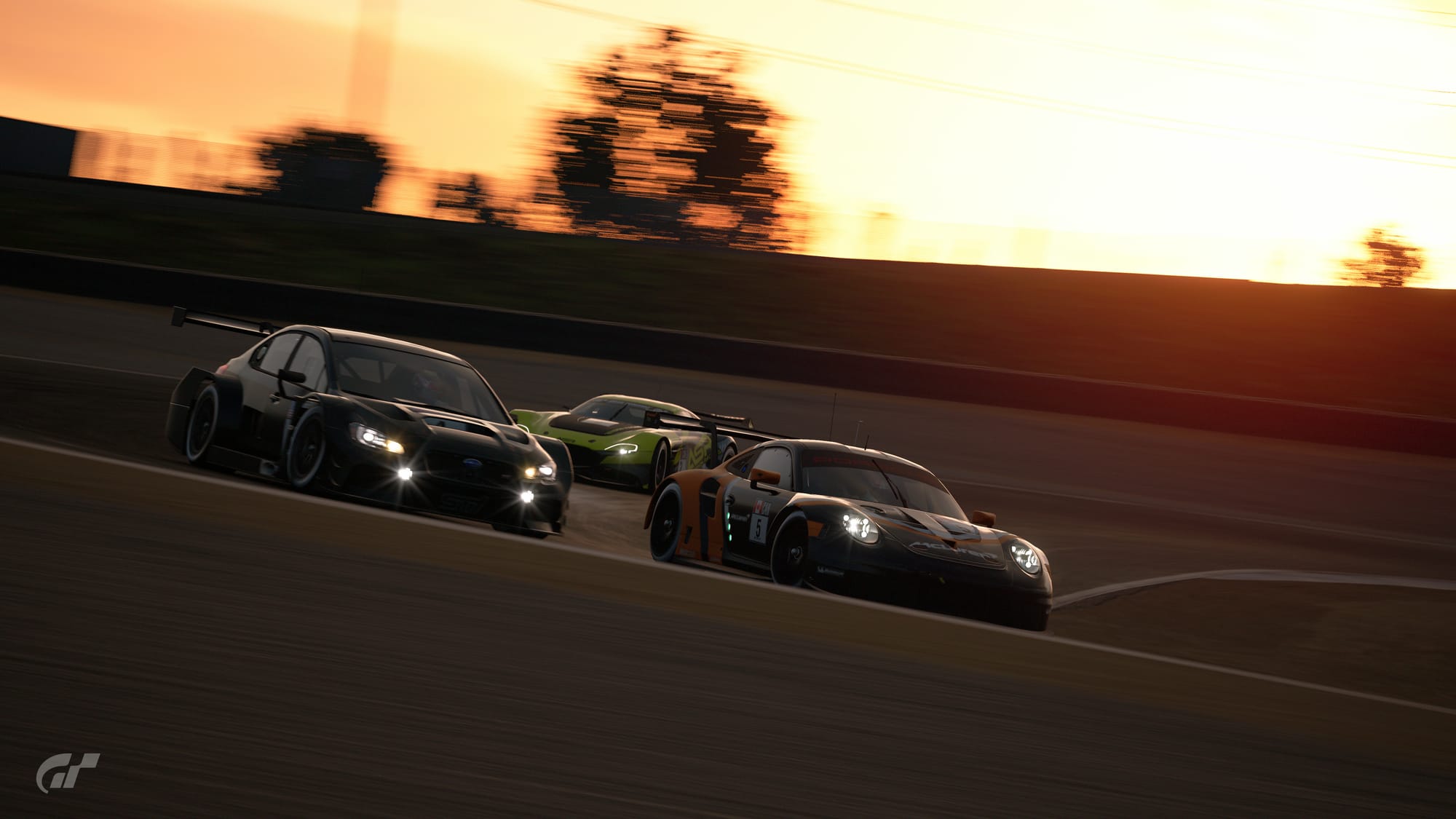 A black with orange McLaren-branded Porsche 911 RSR goes through the Andretti Hairpin at Laguna Seca Raceway. It is side by side with a all-black Subaru WRX Gr.3.