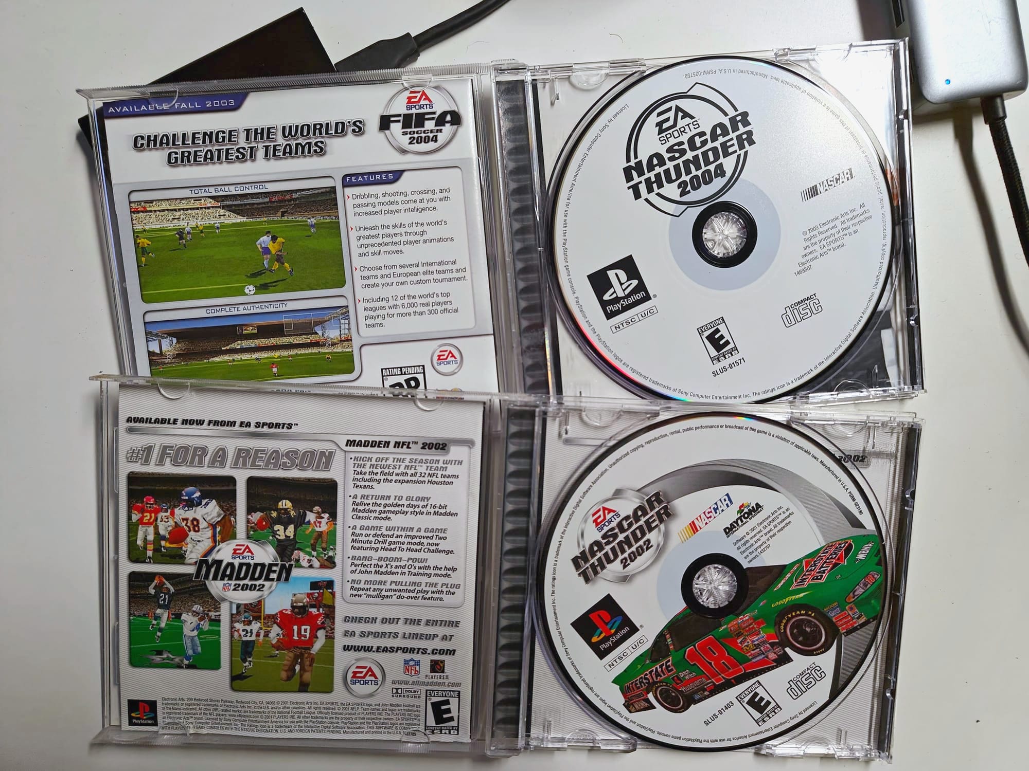 The discs cases for NASCAR Thunder 2002 and 2004 opened up on a white coffee table, with a external hard drive behind. The differences between the two discs is apparent, the 2002 disc has color and features Bobby Labonte's Pontiac Grand Prix on it, while the 2004 disc is non-descript and only in black and white.