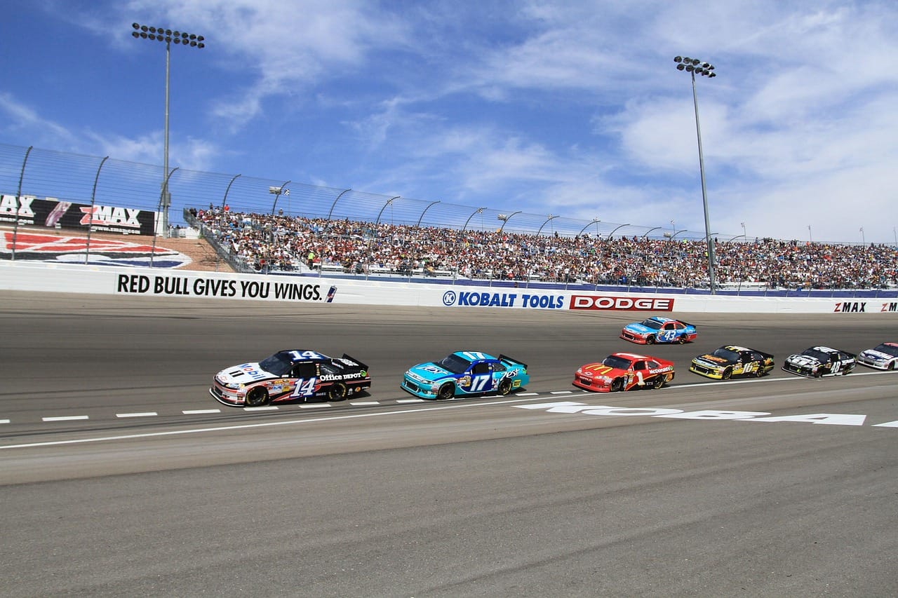 A line of six NASCAR Cup Cars go down off the race track to enter the pit lane at Las Vegas Motor Speedway. They are being led by Tony Stewart in a black with white Mobil 1 Chevrolet.