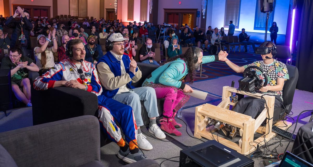 A rally co-driver in a teal and pink drivers suit giving a handshake to Johaan driving on a wooden sim racing rig while wearing a VR headset.