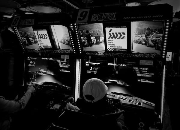 A black and white photo of two SEGA World Drivers Championship arcade racing cabinets in use. The left cabinet is number eight, the right number nine..
